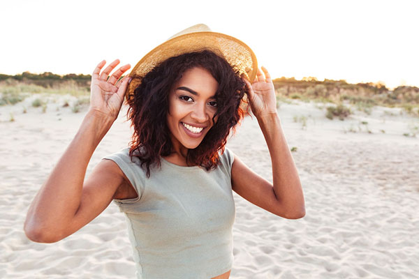 young woman smiling at the beach while holding her sun hat